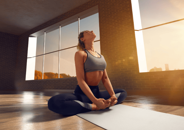 A person sitting on a yoga mat in a bright room, stretching with their face lifted toward the sunlight.