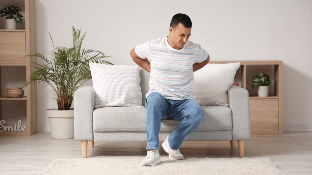 A man sitting on a sofa with his hands on his lower back, appearing to be in pain