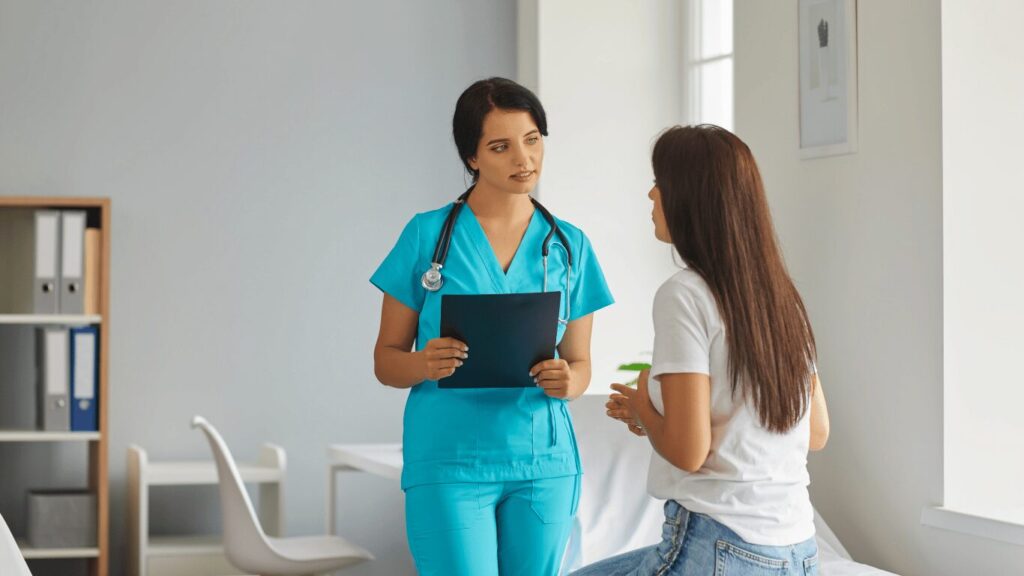 A healthcare professional in blue scrubs holds a clipboard and talks to a patient seated on a bed in a medical office