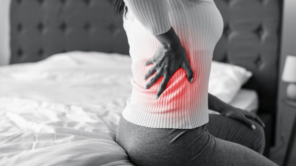 A person is sitting on a bed with their hand on their lower back, where a red highlight indicates pain