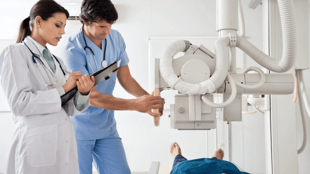 Two medical professionals, one in a white lab coat and the other in blue scrubs, stand near an X-ray machine with a patient lying on a table underneath